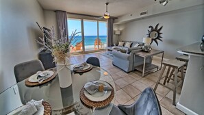 A modern living room with a sofa, dining table, and chairs. There is a large window with a view of the ocean.