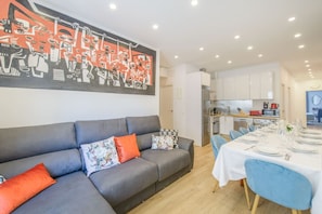 A modern living room with a grey sofa, orange and patterned cushions, a dining table set for four, and a kitchen in the background.