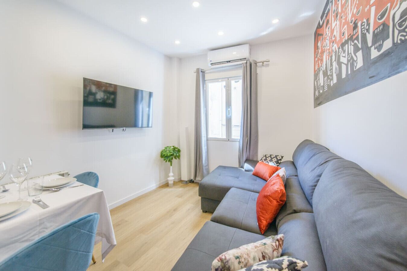 Una sala de estar moderna con un sofá gris, una mesa de comedor con platos y vasos, un televisor de pantalla plana en la pared y un ventanal con cortinas.