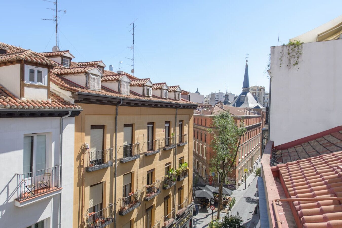 A narrow street with terracotta-tiled buildings, balconies with railings, and a church spire in the background.