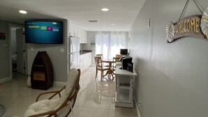 Una sala de estar moderna con un televisor de pantalla plana, una silla de mimbre, un comedor con mesa y sillas, y un cartel que dice 'WELCOME' en la pared.