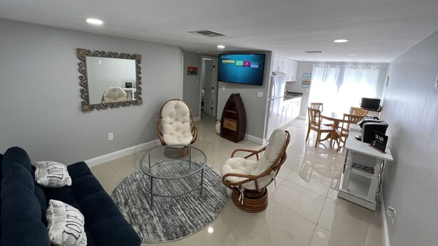 Una sala de estar moderna con un sofá azul oscuro, dos sillas blancas acolchadas, una mesa de centro de vidrio, un televisor de pantalla plana, una zona de cocina con armarios blancos y una mesa de comedor con sillas.