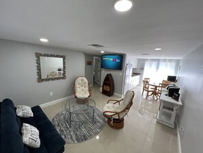 A modern living room with a dark blue sofa, two white cushioned chairs, a glass coffee table, a flat-screen TV, a kitchen area with white cabinets, and a dining table with chairs. - Lovely comfy clean apartment. Close to State parks/ beaches/ restaurants.   (Port Richey)