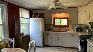 Une cuisine équipée d’un réfrigérateur, d’un évier et d’une cuisinière. On y trouve également un ventilateur de plafond, une fenêtre avec des stores et un tapis sur le sol.