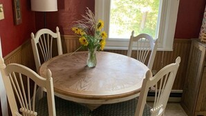 Un coin repas avec une table ronde en bois entourée de quatre chaises blanches, un vase avec des tournesols et une fenêtre donnant sur des arbres.