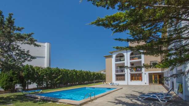 Ein Pool mit klarem, blauem Wasser, umgeben von einer Rasenfläche und Bäumen. Liegestühle und ein kleines Gebäude mit Balkon sind ebenfalls vorhanden.