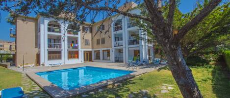 A multi-story building with a swimming pool, outdoor seating, and a tree in the foreground.