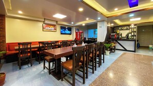 A restaurant with wooden tables and chairs, a television mounted on the wall, a Coca-Cola refrigerator, and a bar area with a piano. - Hotel Abi International (Kathmandu)