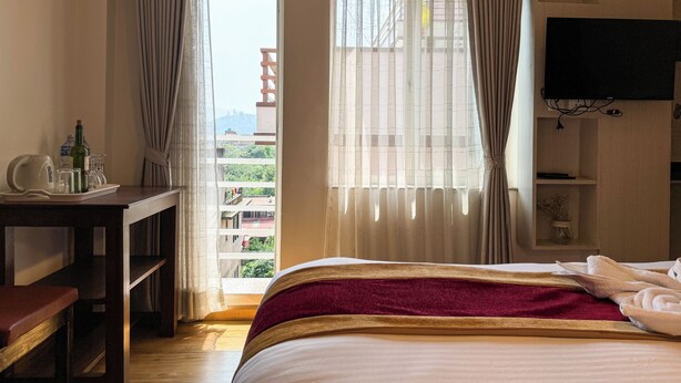 A hotel room with a bed, a desk, a TV, and a view of a building through the window. - Hotel Abi International (Kathmandu)