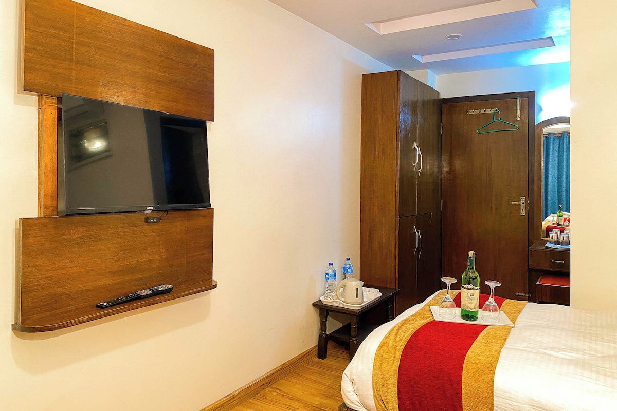 A hotel room with a flat-screen TV mounted on the wall, a wooden wardrobe, a bed with a red and gold bedspread, and a small table with a tray holding a bottle and glasses.