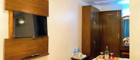 Habitación de hotel con televisor de pantalla plana montado en la pared, armario de madera, cama con colcha roja y dorada, y una mesita con bandeja que contiene una botella y vasos.