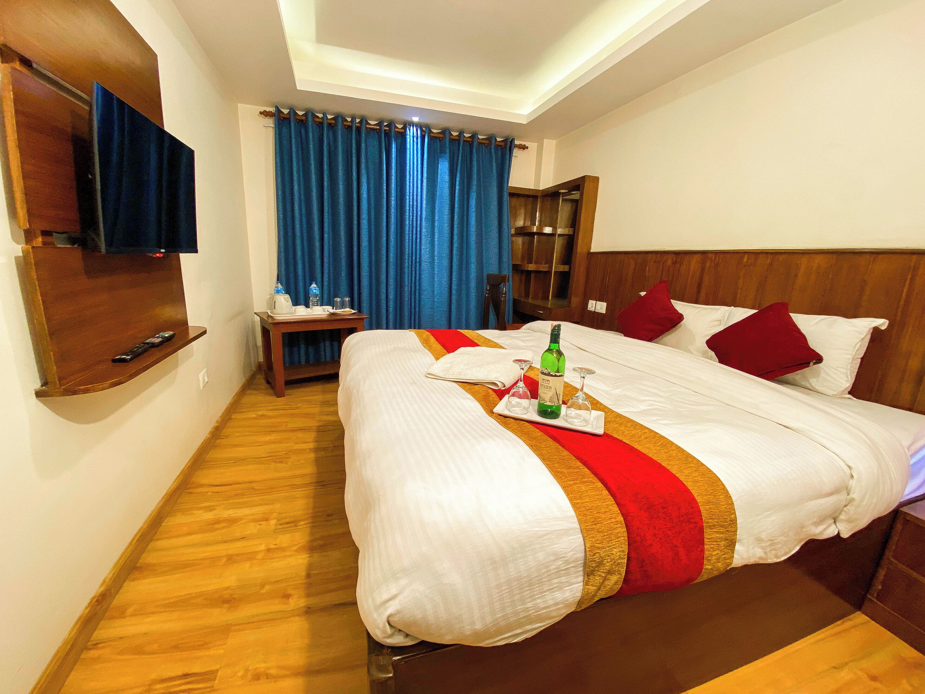 A hotel room with a large bed, a TV mounted on the wall, a wooden nightstand, and a window with blue curtains.