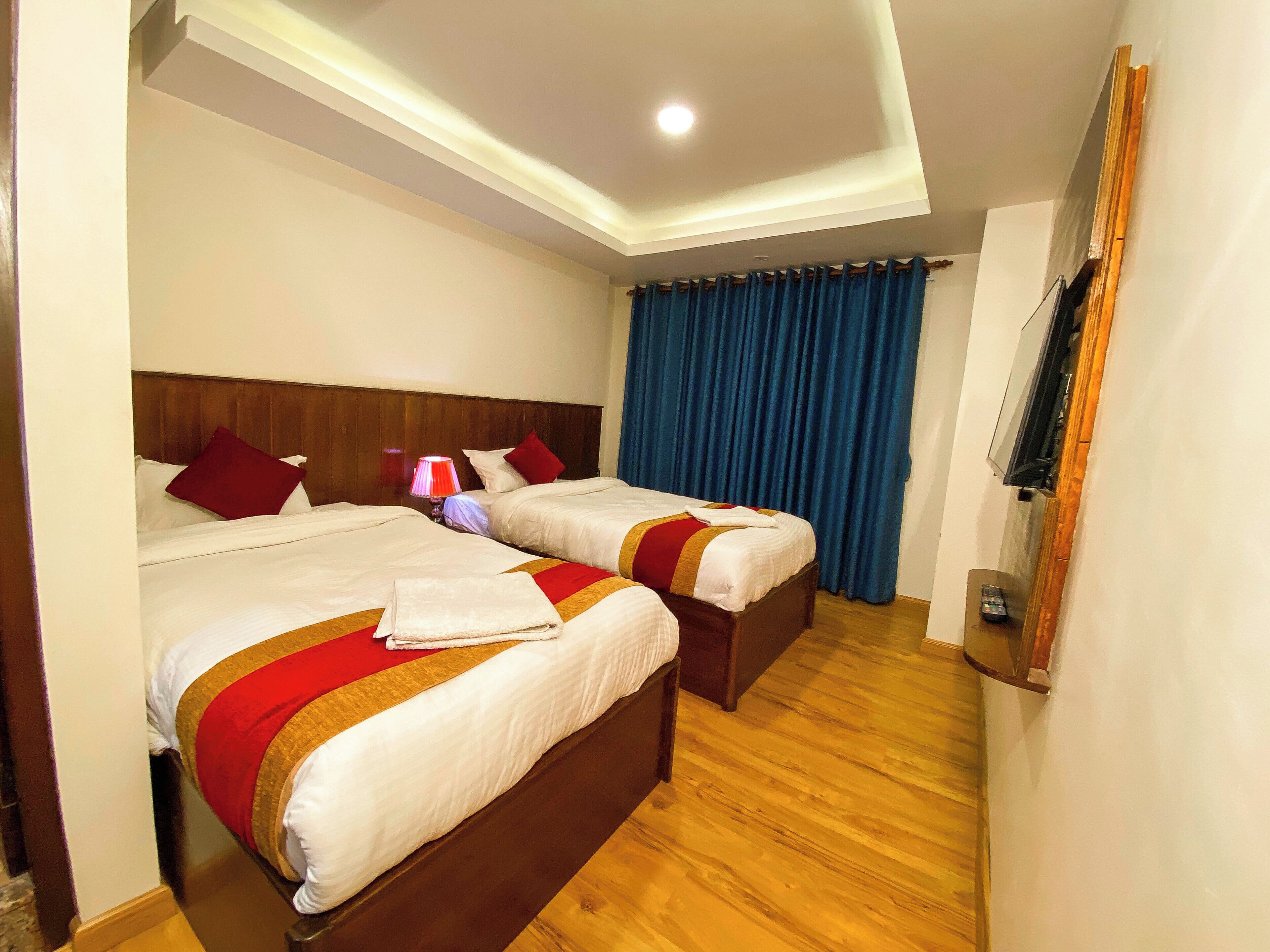 Ein Hotelzimmer mit zwei Betten, Holzboden, einer Deckenleuchte, einem an der Wand montierten Fernseher und einem blauen Vorhang.