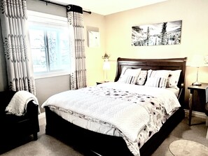 A bedroom with a large bed, a nightstand, a lamp, and a window with curtains. - Legal Walkout suite with King-bed, Kitchen, Parking & Patio (Calgary)