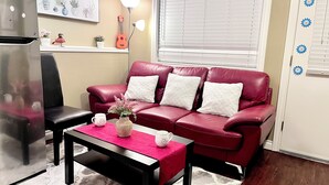 Un salon avec un canapé en cuir rouge, un fauteuil noir, une table basse avec un vase, un réfrigérateur, une lampe et une fenêtre avec des stores.