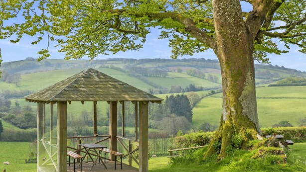 A wooden gazebo with a roof, benches, and a table, surrounded by lush green fields and a large tree.