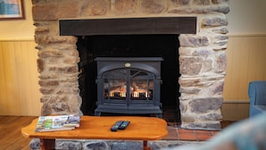 A stone fireplace with a black mantel, a wooden table with magazines and remotes, and a blue chair. - Stable Cottage near Wheddon Cross (Minehead)