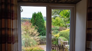 A room with a large window offering a view of a garden with various plants and a tall tree. - Stable Cottage near Wheddon Cross (Minehead)