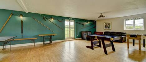 A spacious room with wooden flooring, a billiards table, a foosball table, and a pool table.