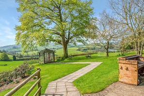 A grassy pathway leading to a gazebo, a brick barbecue area, and a picnic table in a rural setting with rolling hills in the background.