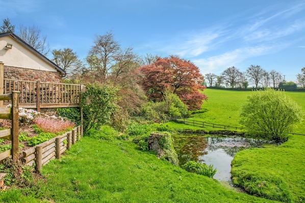 A house with a stone facade, a wooden deck, and a small pond in a lush green landscape.