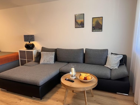 Ett modernt vardagsrum med en hörnsoffa, ett litet träbord med ett ljus och frukt på, och två reseaffischer på väggen.