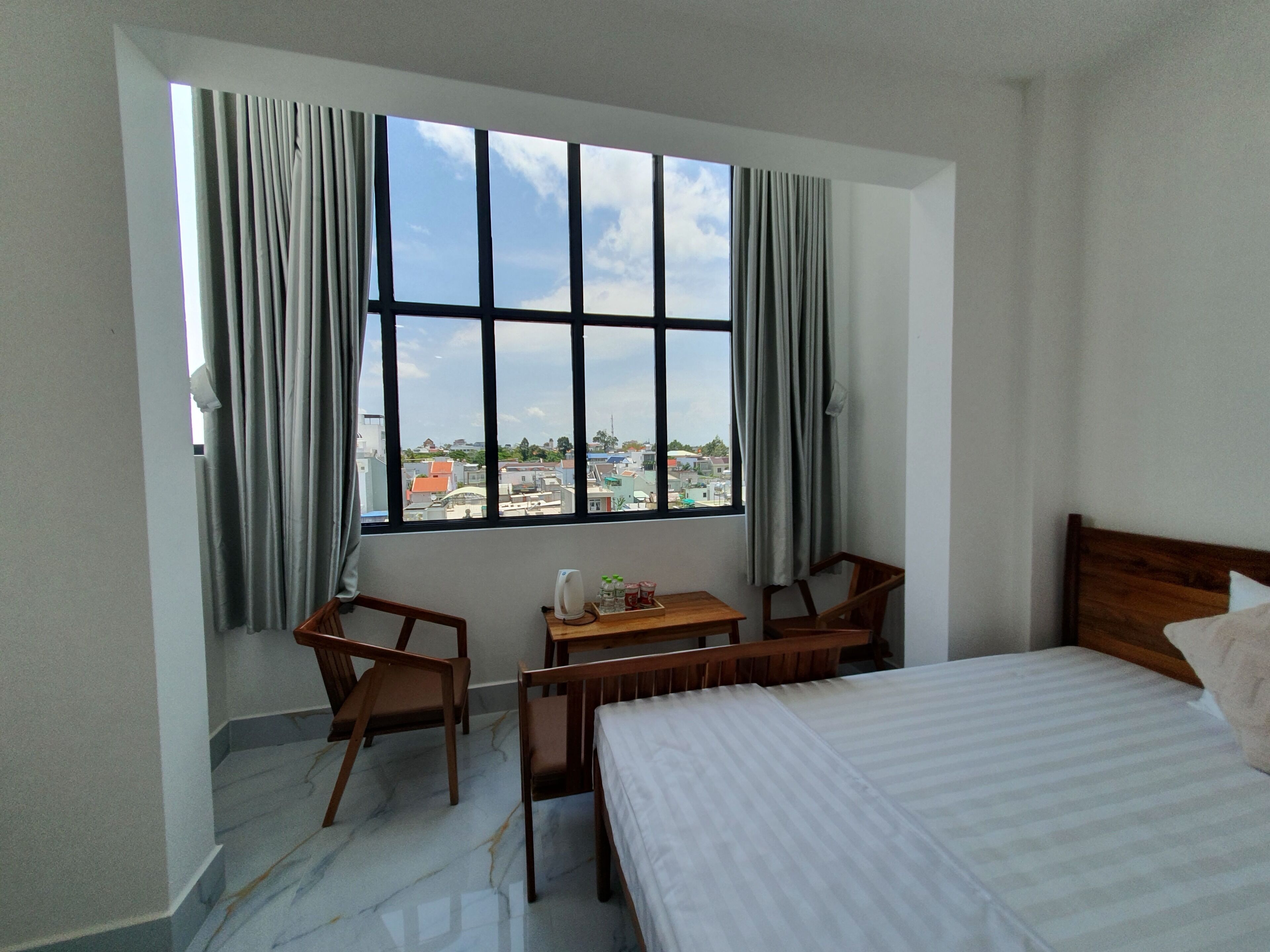 A hotel room with a bed, a desk with chairs, a view of the city through large windows, and a view of the sky.