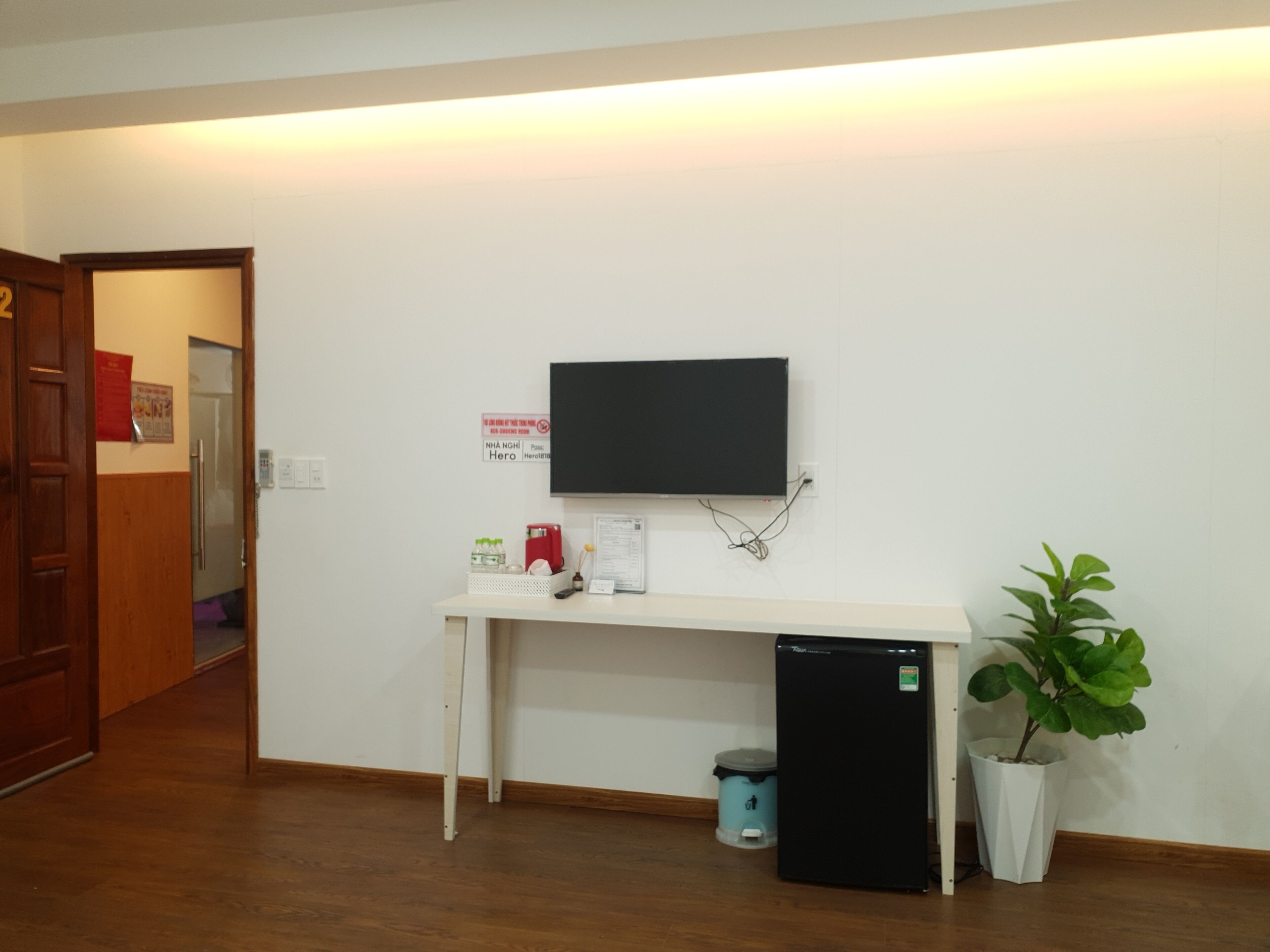 A room with a white desk, a black TV mounted on the wall, a small black refrigerator, a trash bin, and a potted plant.