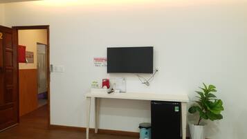 A room with a white desk, a black TV mounted on the wall, a small black refrigerator, a trash bin, and a potted plant.