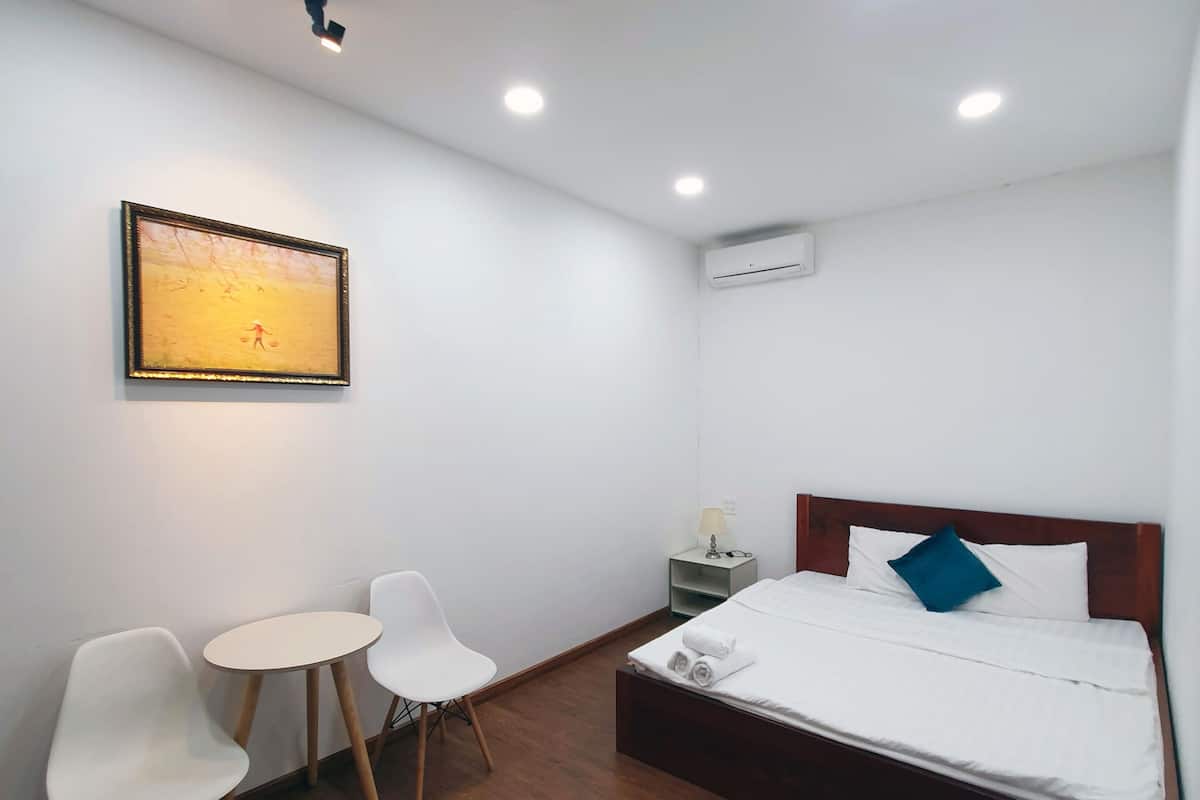 A hotel room with a bed, two white chairs, a small table, a painting on the wall, and recessed ceiling lights.