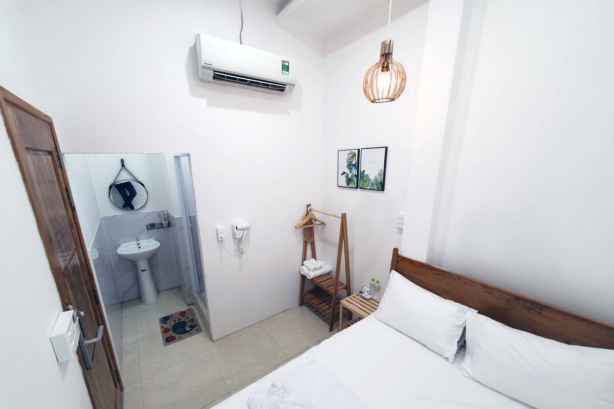 A hotel room with a bed, a bedside table, a hanging light fixture, a wall-mounted air conditioner, a mirror, a sink, and a towel rack.
