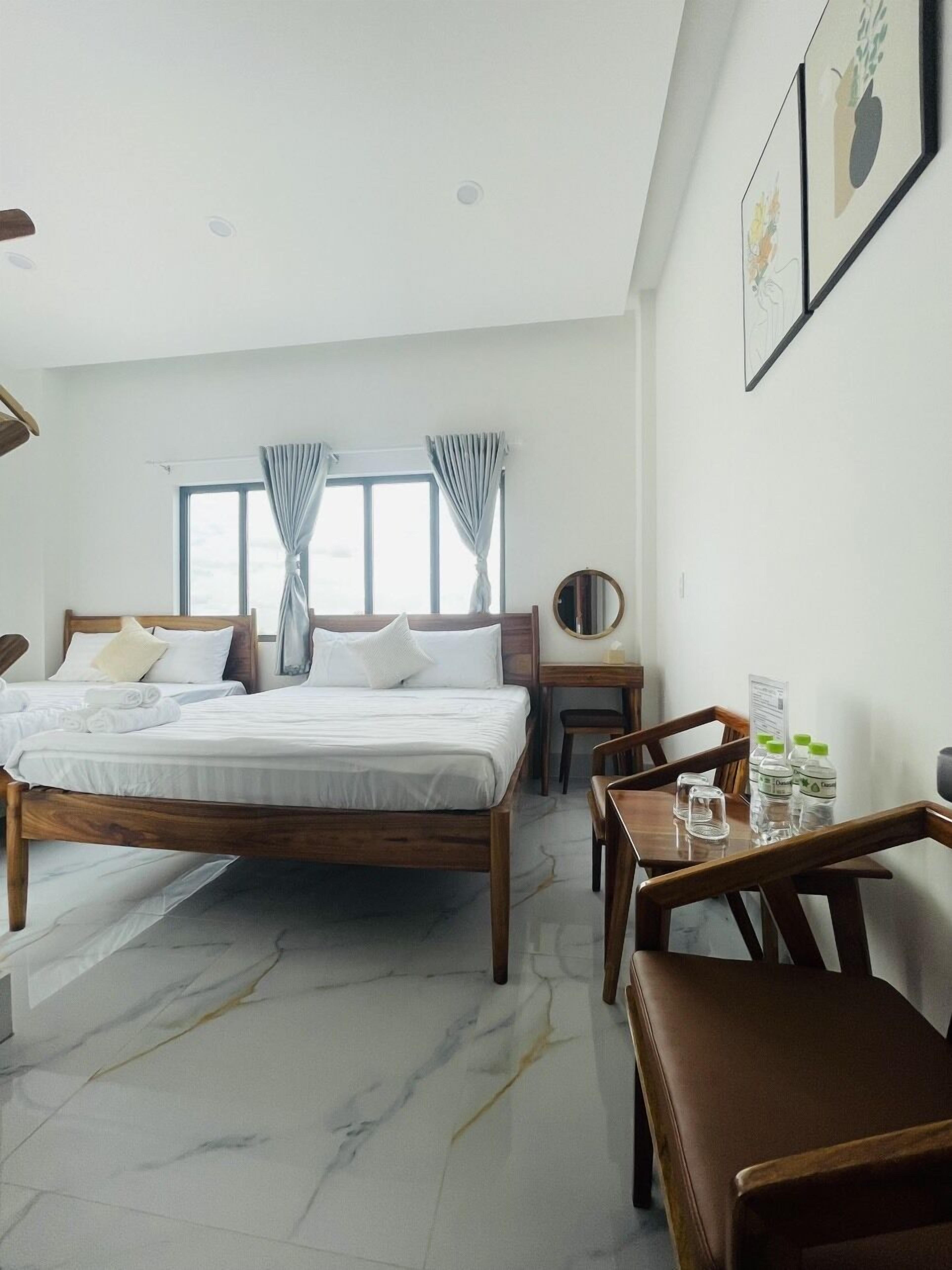 Une chambre d’hôtel avec un grand lit, un bureau en bois et une chaise. Les fenêtres sont dotées de rideaux, et un miroir rond est accroché au mur.