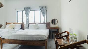 Une chambre d’hôtel avec un grand lit, un bureau en bois et une chaise. Les fenêtres sont dotées de rideaux, et un miroir rond est accroché au mur.