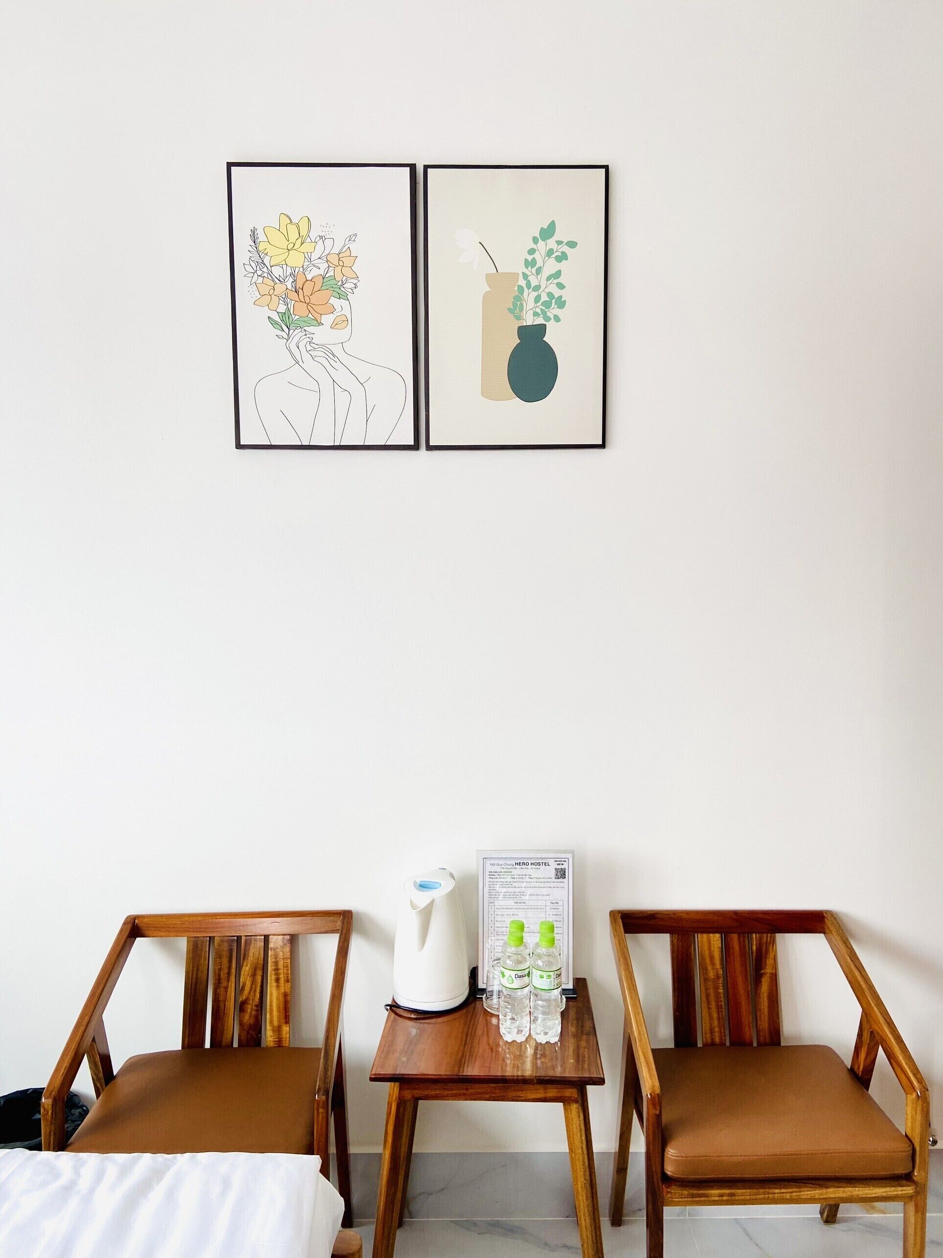 Una habitación con dos sillas de madera, una mesita con hervidor y agua embotellada, y dos cuadros enmarcados en la pared.