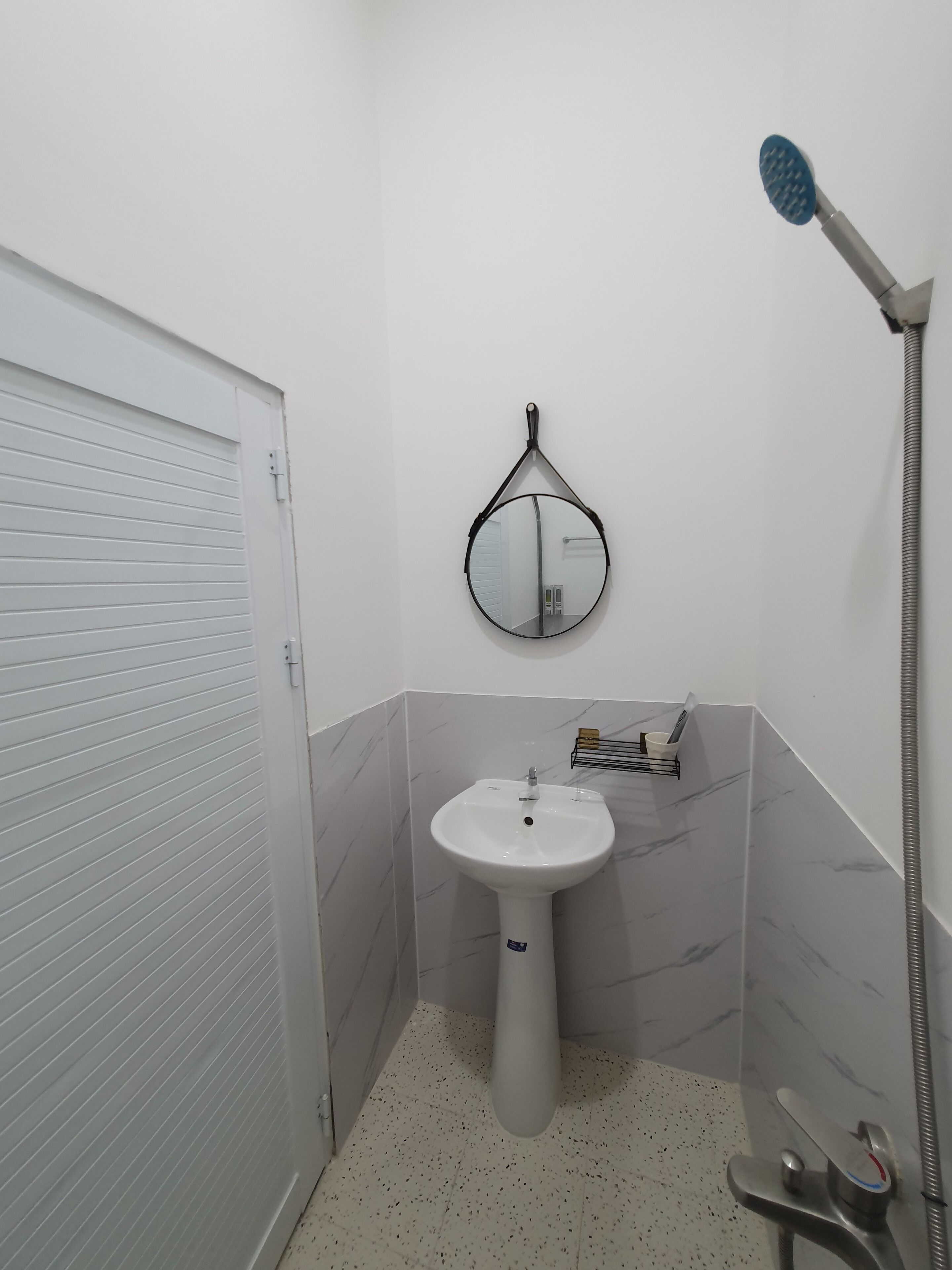 A bathroom with a white tiled floor, a pedestal sink, a round mirror, and a handheld showerhead.