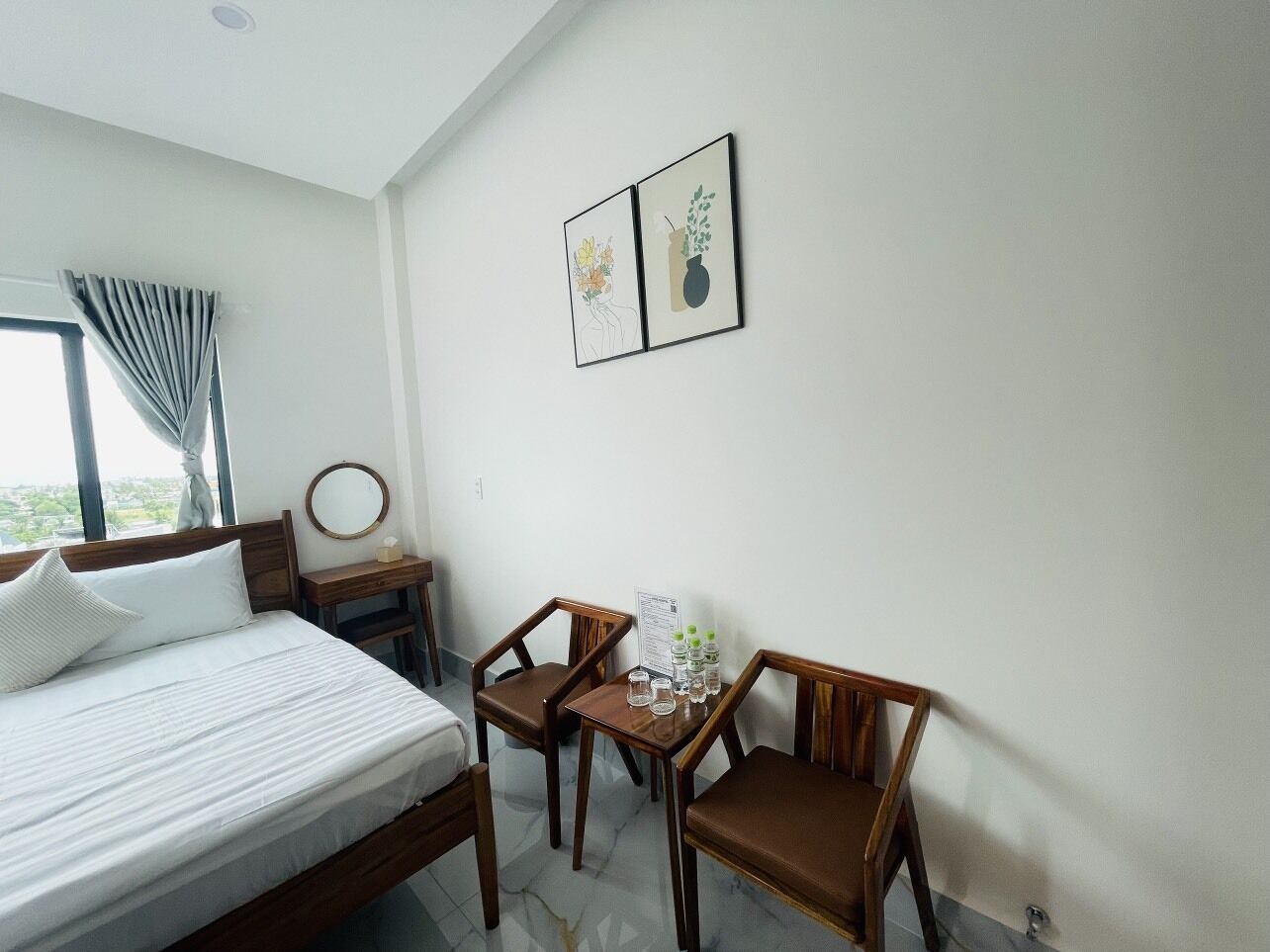 Habitación de hotel con una cama, dos sillas de madera, una mesita con botellas de agua y dos cuadros en la pared.