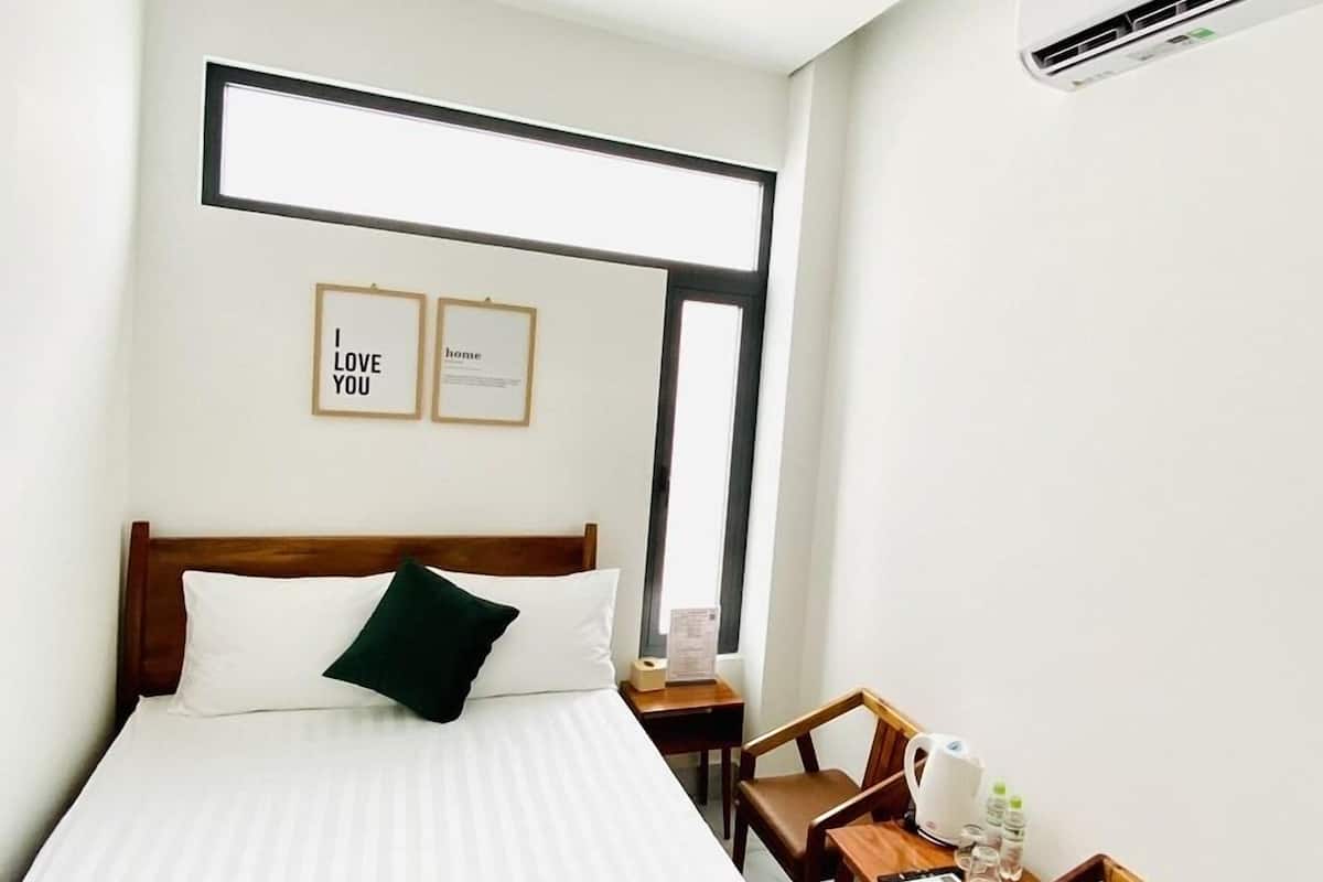 A neatly made bed with white linens and a dark pillow, a small wooden table with a kettle and glasses, and two framed pictures on the wall.