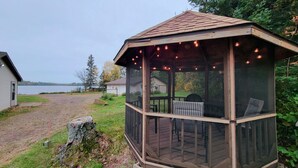 Un belvédère en bois doté d’un espace salon couvert, offrant une vue sur un lac et entouré d’arbres.