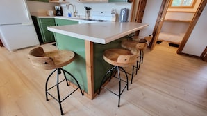 Une cuisine avec un plan de travail blanc, des armoires vertes et des tabourets en bois aux pieds métalliques.