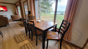 Un coin repas avec une table et des chaises en bois, de grandes fenêtres avec des rideaux, une cheminée et une vue sur un lac.