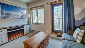 Een woonkamer met een grote flatscreen televisie, een open haard, een houten bankje, een bank en een raam met gordijnen.