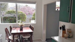Un área de comedor con una mesa y sillas de madera, una ventana con vistas a árboles y una cerca, y una cocina verde con armarios y una encimera.