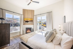 Un dormitorio con una cama grande, una chimenea, un ventilador de techo y vistas a un paisaje nevado a través de las ventanas.