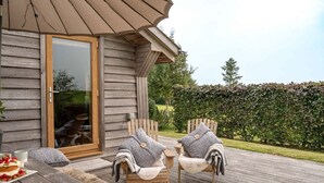 Una terraza de madera con dos sillas y una mesa, una puerta de madera y vistas a un seto y árboles.