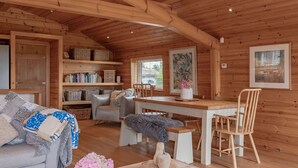 Un acogedor interior de cabaña con techo de madera, una mesa de comedor con sillas, un sofá y estanterías con libros y cestas.