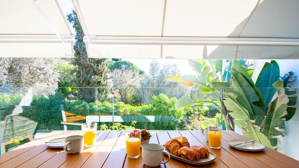 A wooden table with breakfast items including croissants, juice, and a coffee cup, set against a backdrop of lush greenery and a clear view of the outdoors. - Casa Ella - Beautifully renovated luxury townhouse walking distance of Carvoeiro (Carvoeiro)