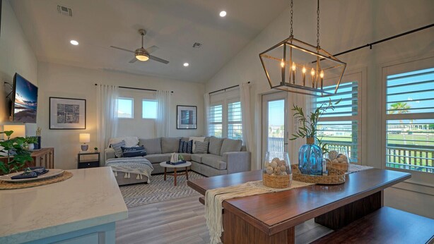 A spacious living area with a large sofa, a coffee table, and a wooden dining table. There is a ceiling fan, a chandelier, and a view of the outdoors through large windows. - New Build! Priv Pool, Firepit, Walk to the Beach | Alright, Alright by AvantStay (Port Aransas)