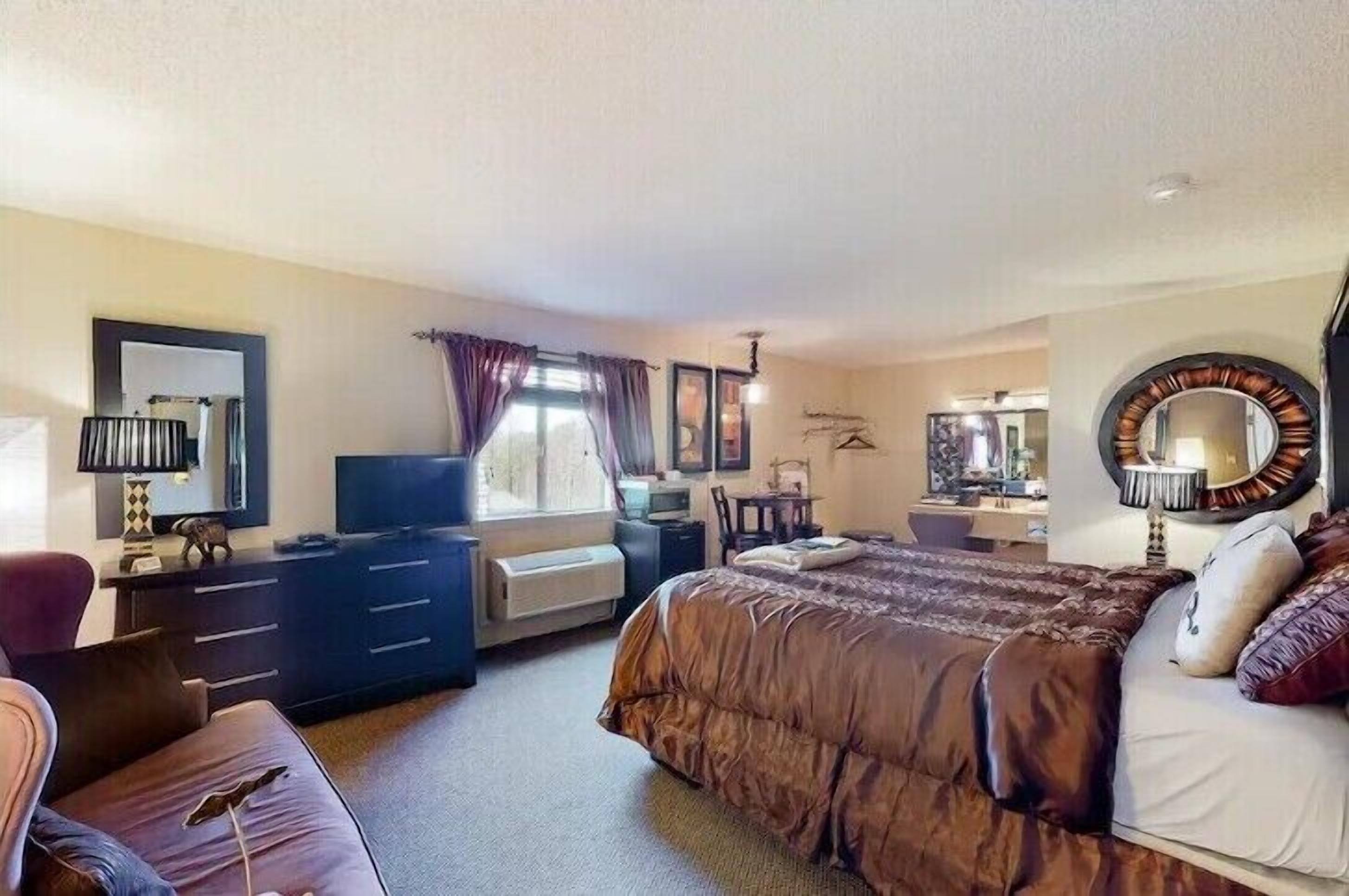 A hotel room with a large bed, a desk with a chair, a television, a dresser, and a window with curtains.