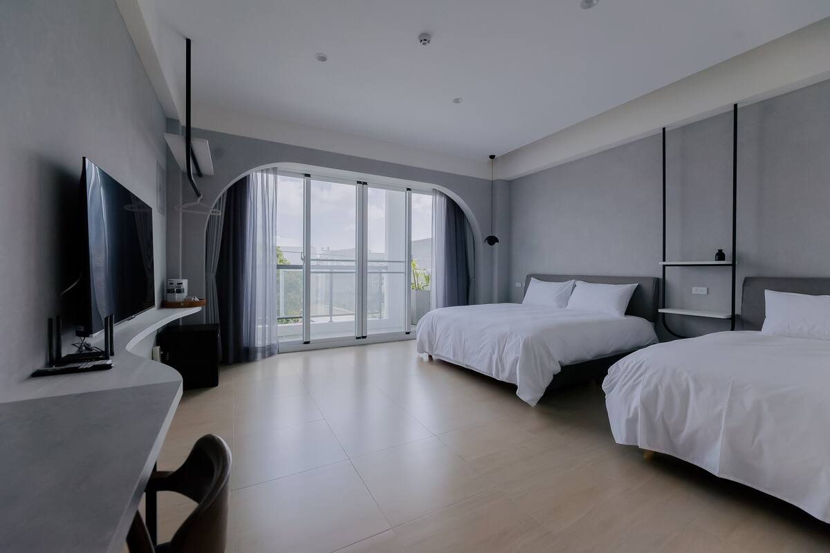 A modern hotel room with two beds, a desk with a computer, and a large sliding glass door to a balcony.