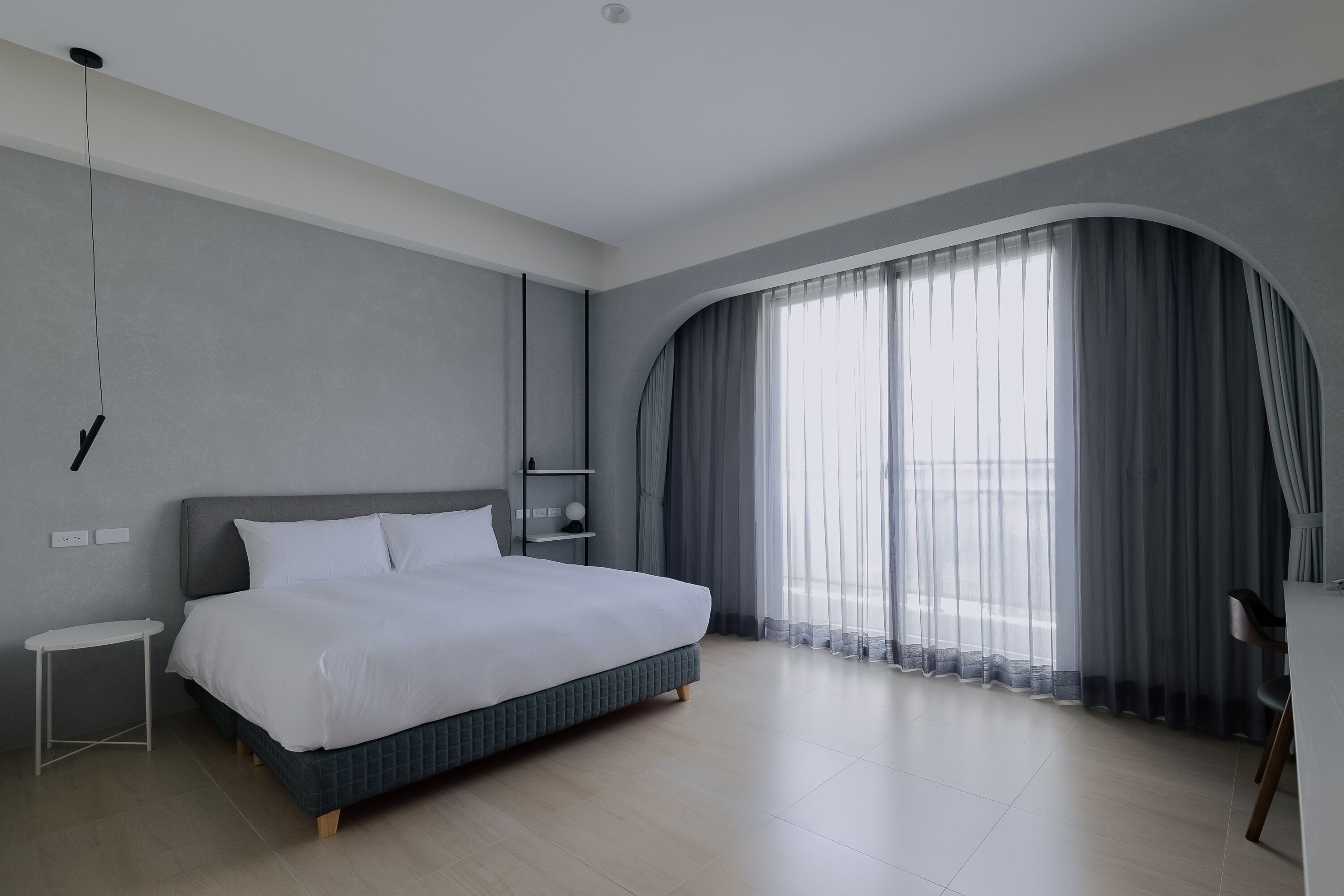 Une chambre moderne avec un grand lit, une petite table de chevet, un bureau et une fenêtre incurvée agrémentée de rideaux légers.
